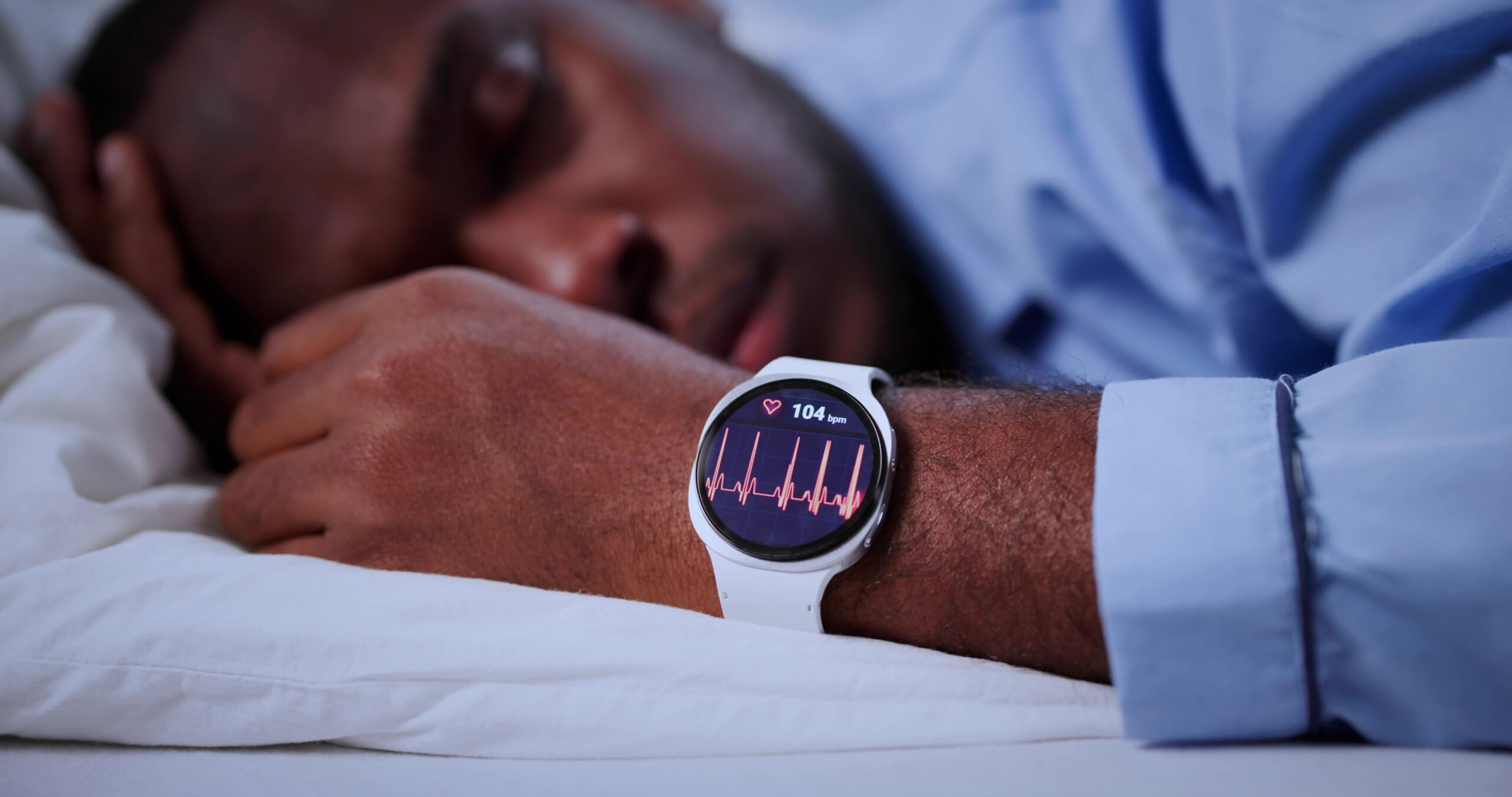 Wrist with a white smartwatch showing heart rate 104 bpm and ECG lines as a person rests in bed.