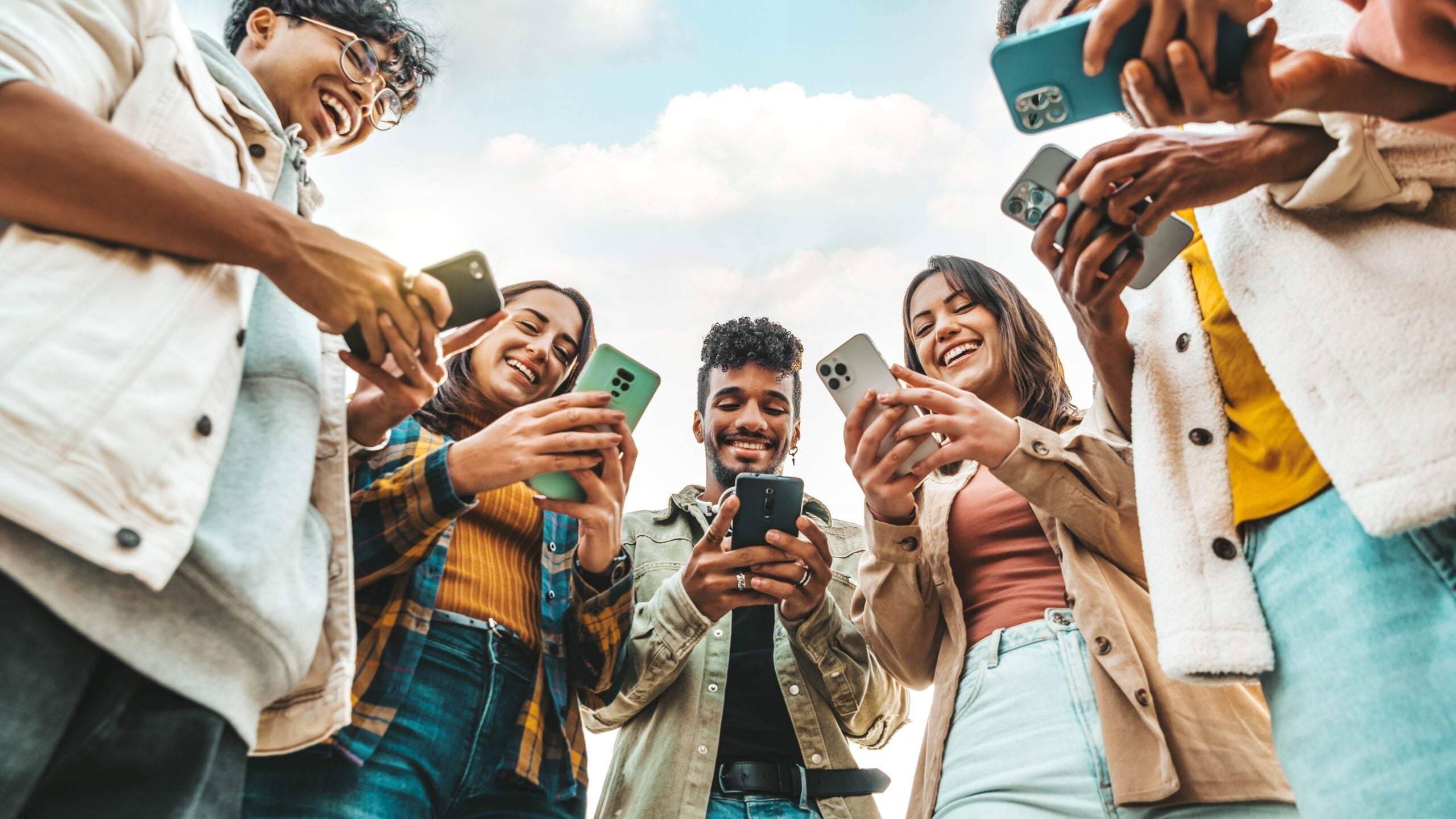 Diverse group of friends standing in a circle, holding smartphones and smiling at the camera.